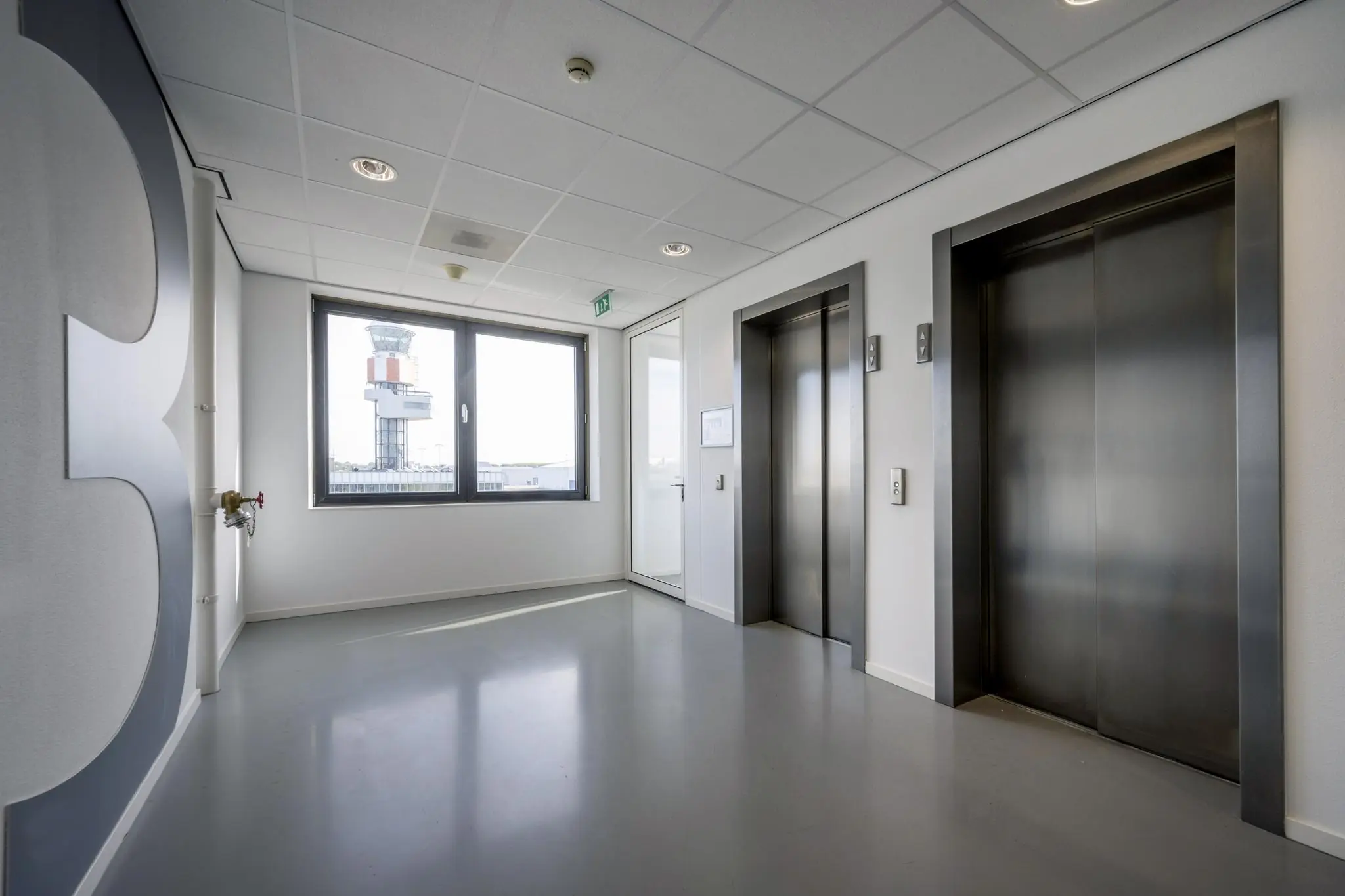 Hal met liften op de derde verdieping van een kantoorpand aan het Rotterdam Airportplein, met zicht op de verkeerstoren van Rotterdam The Hague Airport.